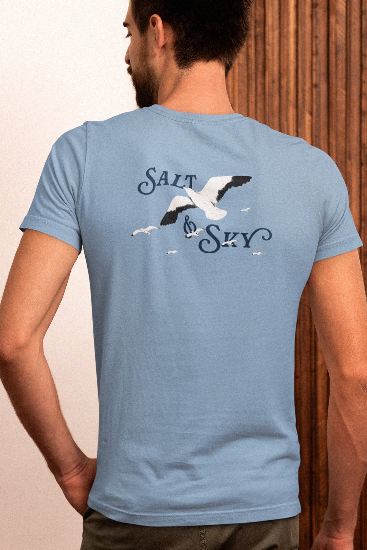 Man wearing a light blue t-shirt with 'Salt & Sky' design on the back, standing against a wooden panel background.